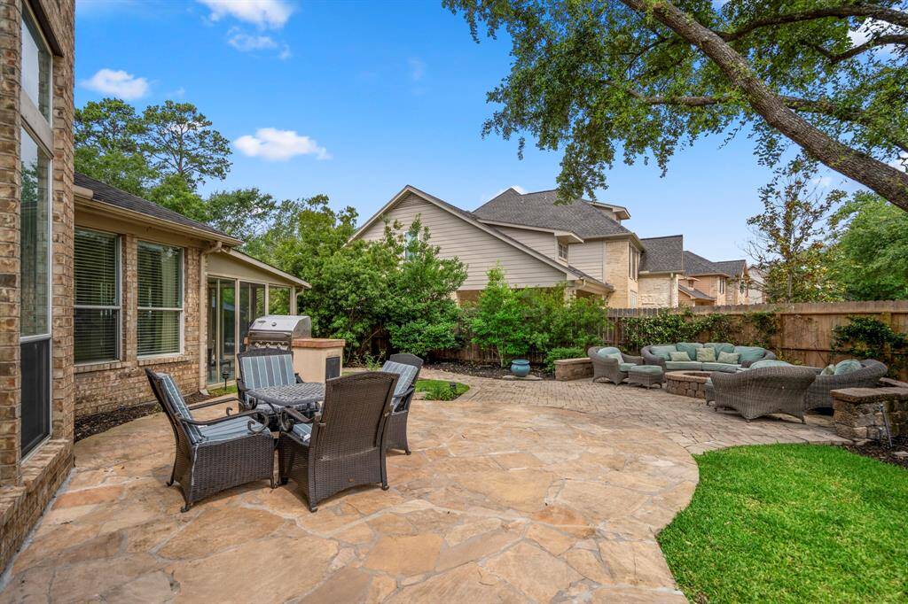This backyard features a spacious stone patio with outdoor seating, a barbecue grill, and lush landscaping. Ideal for entertaining and relaxation, it offers a private and serene atmosphere.