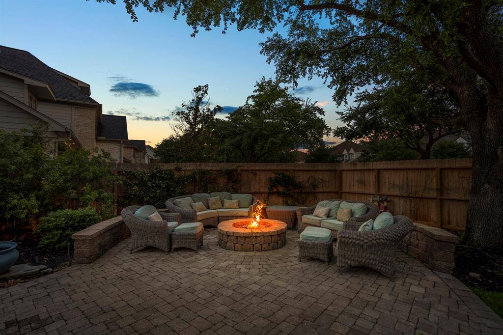 This inviting backyard features a cozy, circular stone fire pit surrounded by comfortable wicker seating. Set on a spacious stone patio, it's perfect for outdoor gatherings and relaxation, all framed by a wooden fence and mature trees for added privacy.