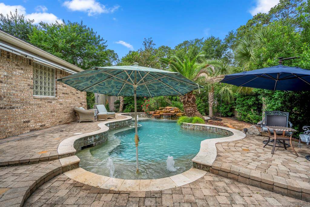 This photo showcases a luxurious backyard with a pool complete with a waterfall feature. It includes a spacious patio area with lounge chairs and umbrellas, surrounded by lush greenery and palm trees for a private, tropical feel.