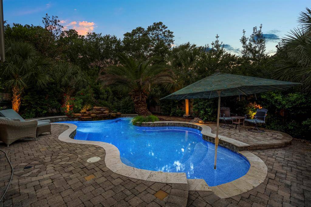 This backyard oasis features a beautifully lit pool with a waterfall, surrounded by lush landscaping and mature trees. The stone patio offers a cozy seating area with a large umbrella, perfect for relaxing evenings.