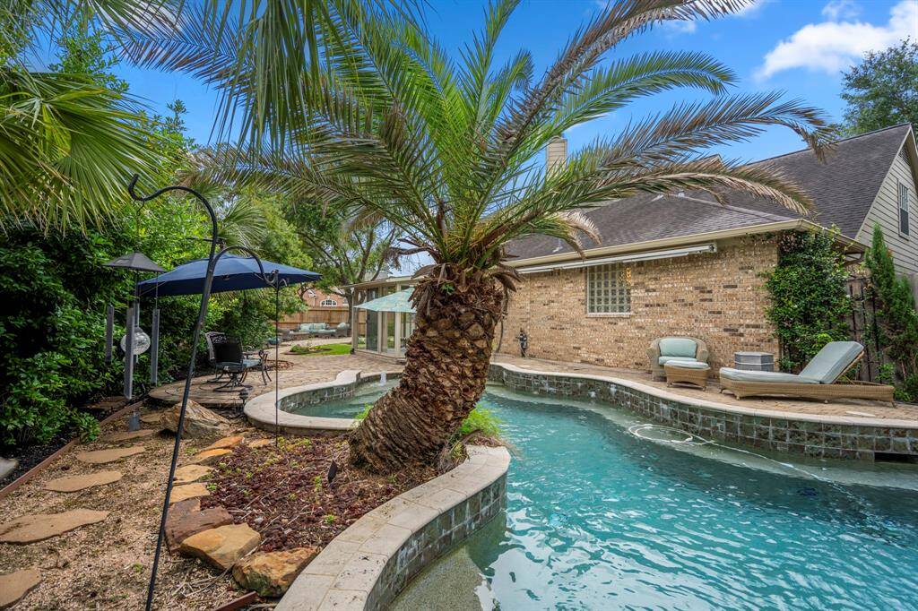 This backyard features a charming pool with a tropical vibe, surrounded by lush greenery and a brick exterior. It includes a cozy seating area with an umbrella and lounge chairs, perfect for relaxation and outdoor enjoyment.