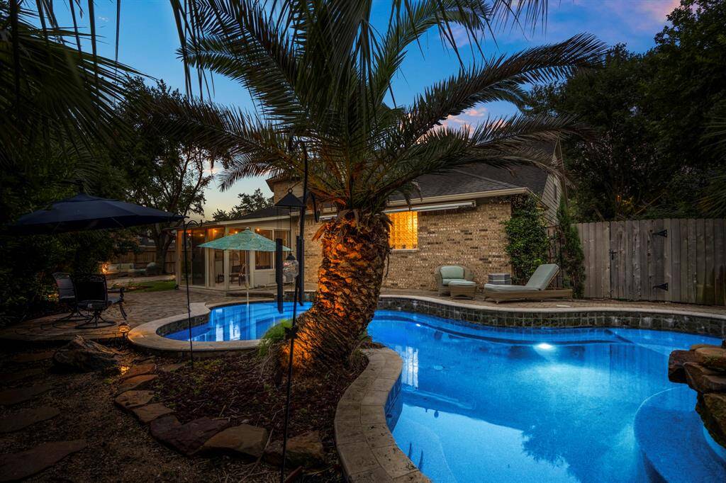 This photo showcases a serene backyard with a beautifully lit pool, surrounded by lush greenery and a cozy seating area, perfect for relaxation and entertaining.