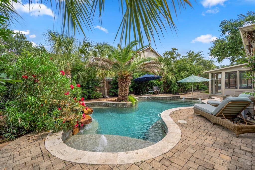 A tropical oasis backyard featuring a pool surrounded by lush greenery and palm trees. The space includes a cozy patio with lounge chairs and umbrellas, perfect for relaxing or entertaining.