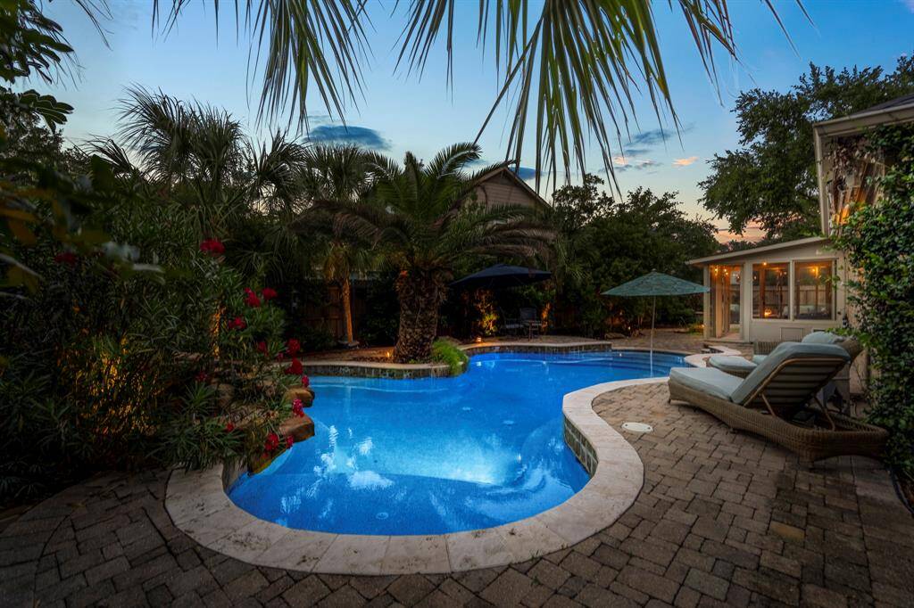 This photo showcases a serene backyard oasis featuring a beautifully lit swimming pool surrounded by lush tropical landscaping. There are comfortable lounge chairs and umbrellas on a paved patio, creating an inviting space for relaxation and entertaining.