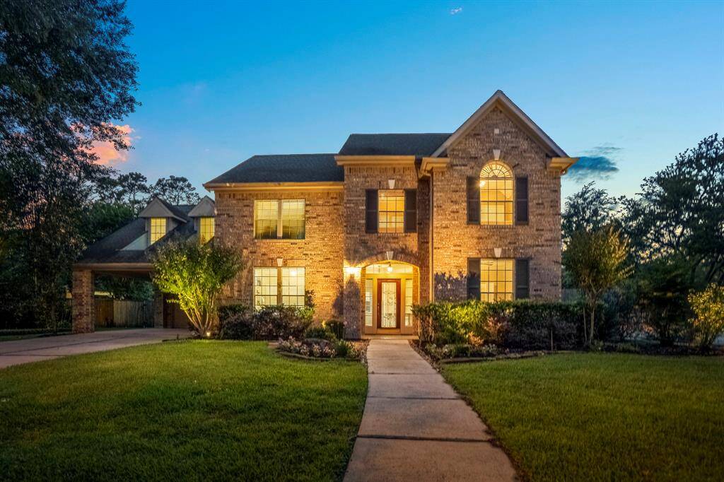 This inviting two-story brick home features a well-maintained lawn, a welcoming front entrance with large windows, and a spacious driveway. The property is beautifully lit, highlighting its charming architecture against the evening sky.