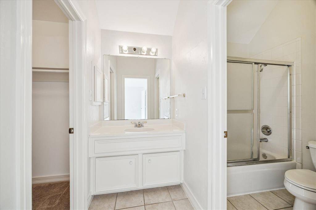 This bathroom features a clean design with a large mirror over a single-sink vanity, a bathtub with sliding glass doors, and an adjacent closet. The space is bright with neutral tile flooring.