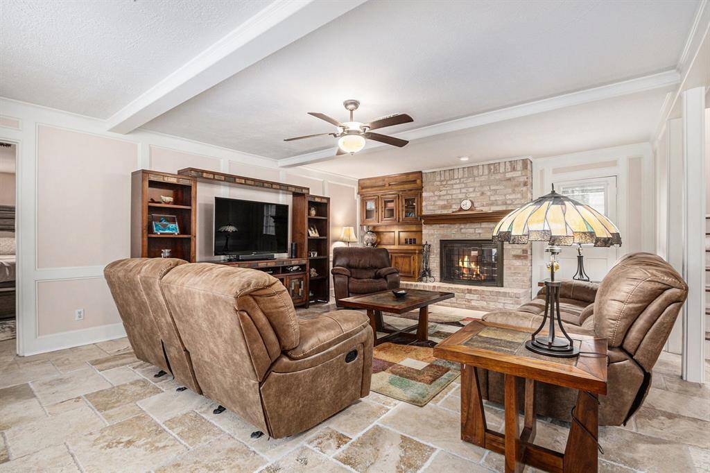 Living area boasts a beamed ceiling, block paneling, crown molding, brick wood burning fireplace flanked by shelving with access to the back patio.