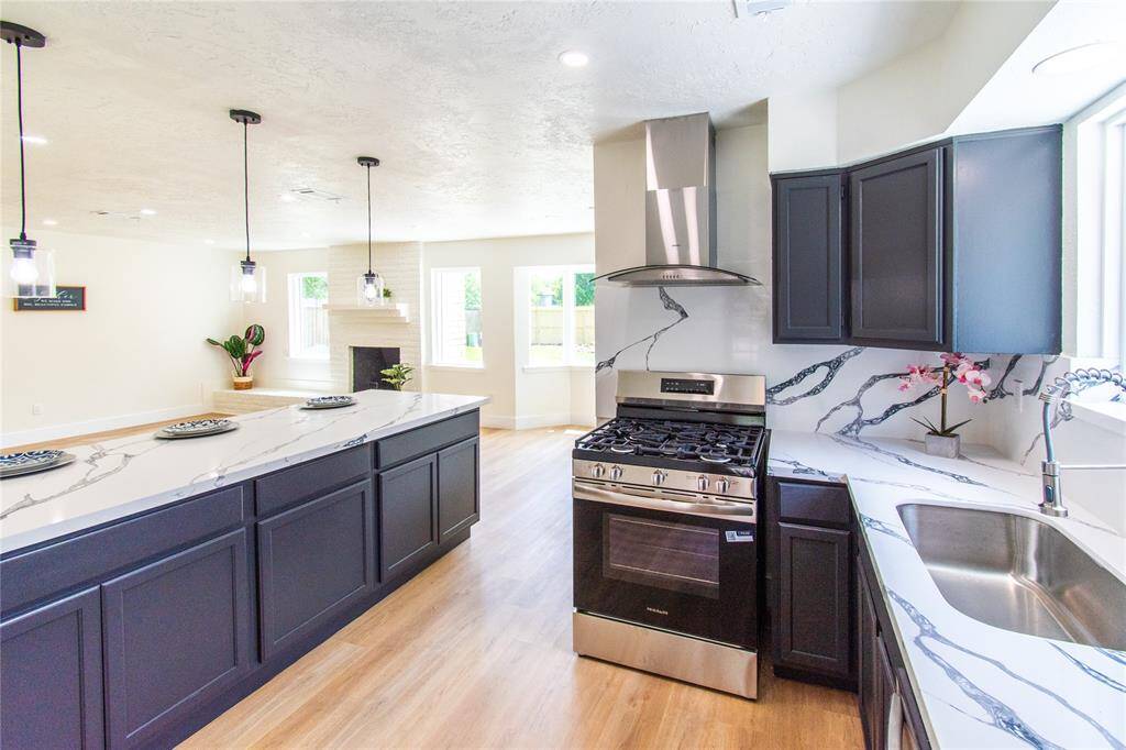 Sleek dark cabinetry, marble-patterned countertops, and stainless steel appliances, including a gas range. The open layout connects to a bright living area with large windows and a cozy fireplace, perfect for entertaining.
