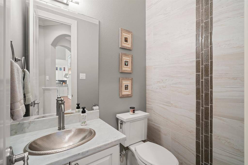 The downstairs guest powder room features tile flooring, crown molding, an artisan-style hammered metal sink with marble countertop, decorative wall tile accents, and toilet.