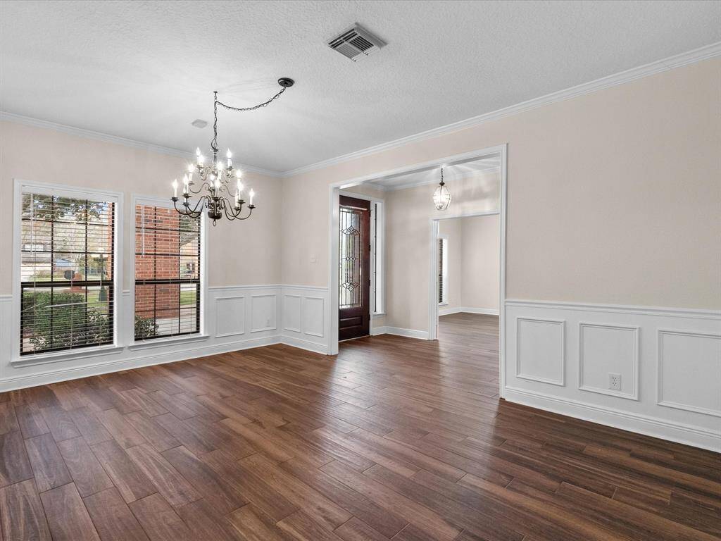 This photo showcases a bright, open dining area with porcelain wood flooring, large windows, and a classic chandelier. The room features elegant wainscoting and opens into an adjoining space, with a view of the front door framed by decorative glass panels.