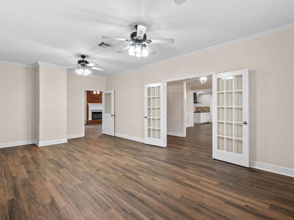 This spacious living/office area features soft beige walls, and two ceiling fans. It includes French doors leading to additional rooms, with a glimpse of a cozy brick fireplace and a modern kitchen, offering a blend of open space and classic charm.