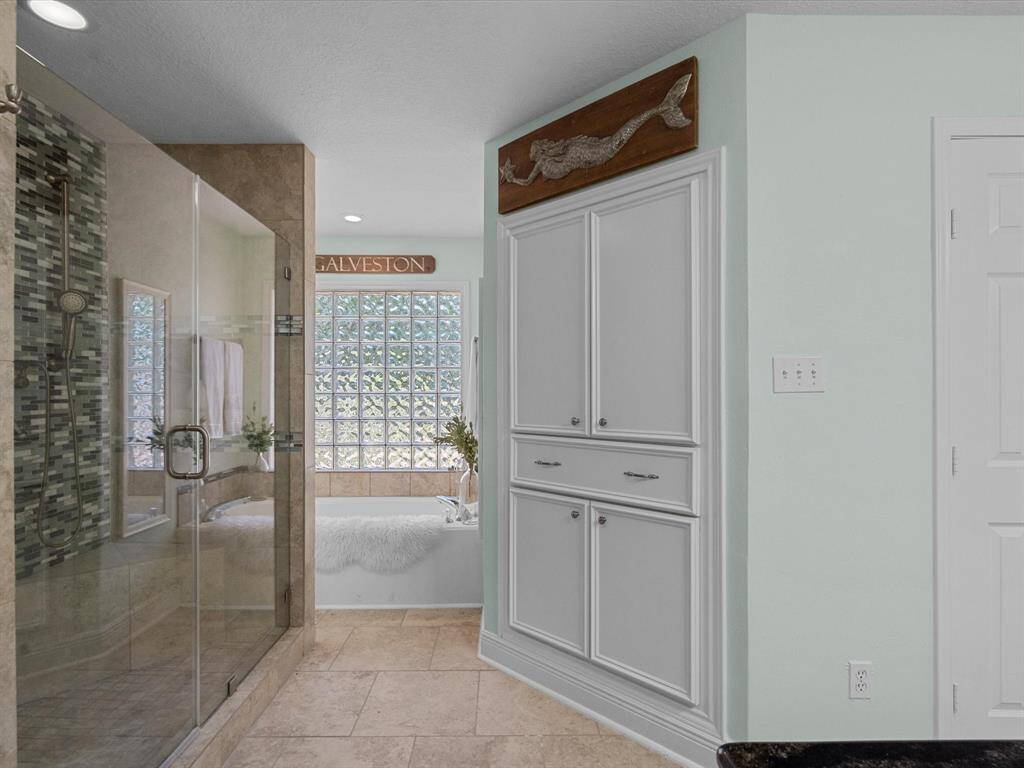 Primary bathroom features a spacious glass-enclosed shower, a built-in bathtub with a glass block window for natural light, ample cabinetry for storage, and a sleek, modern design.