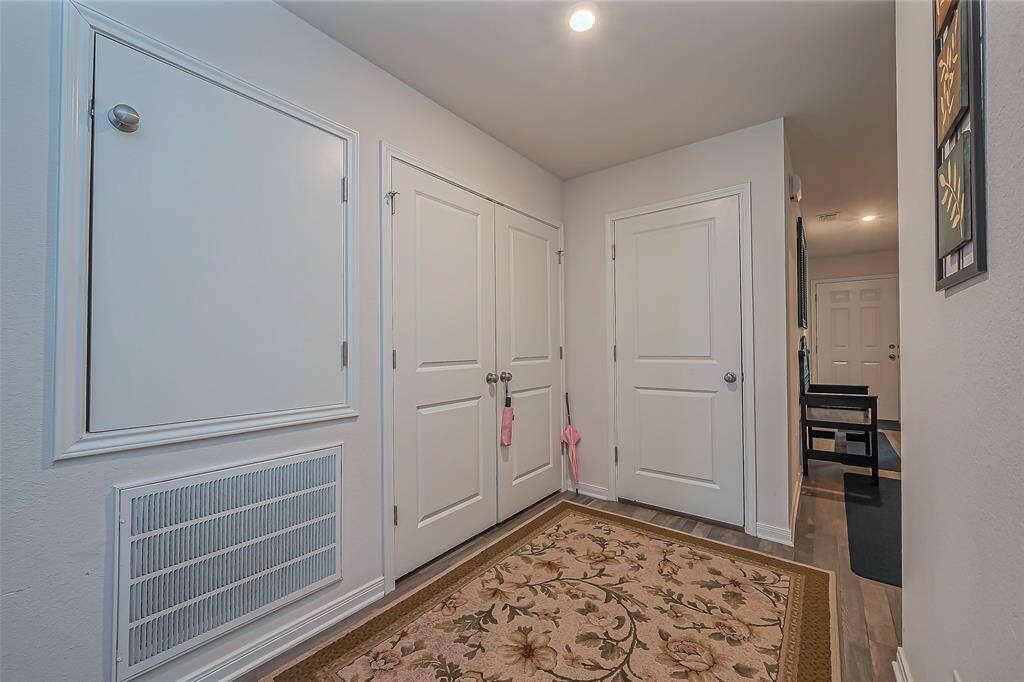 This photo shows a hallway with white walls and doors, featuring ample storage space. The flooring is wood, complemented by a decorative floral rug. Recessed lighting provides a bright, welcoming atmosphere.