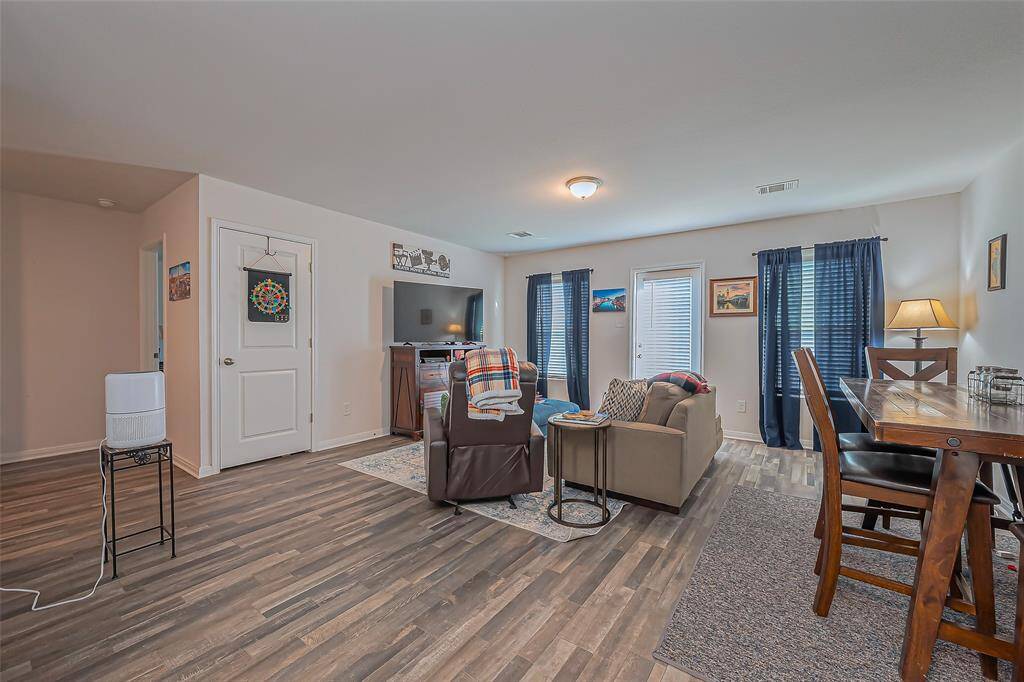 This cozy living space features an open layout with wood-style flooring, a comfortable seating area, and a dining nook. Natural light streams through large windows with blue curtains, and there's a TV setup against the wall. Perfect for relaxing and entertaining.