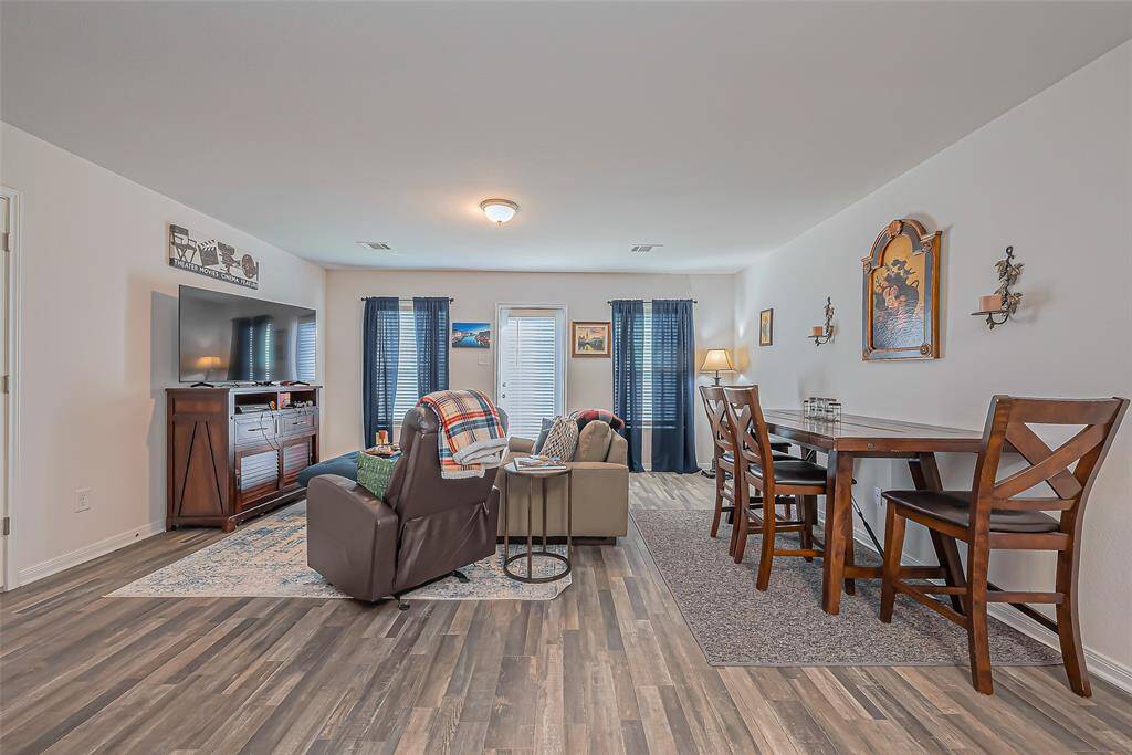 This spacious living area features modern laminate flooring and a cozy setup with a large TV and comfortable seating. It includes a dining area with a stylish wooden table and chairs, accented by natural light from windows with dark curtains.