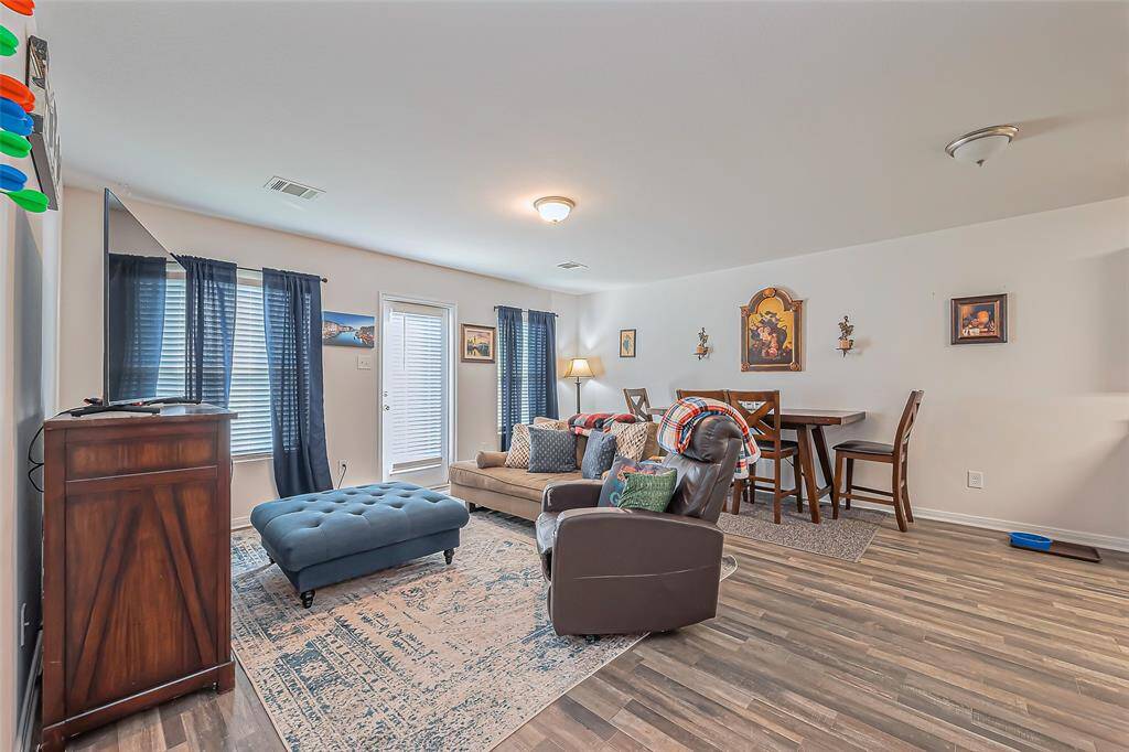 This inviting living area features laminate wood flooring, ample natural light from multiple windows, and a cozy setup with a couch, recliner, and ottoman. The space includes a dining nook with a high table and chairs, complemented by tasteful wall decor.