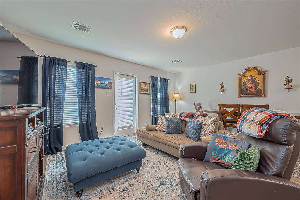 This cozy living room features a comfortable sofa and armchair, accented with soft pillows and throws. Natural light streams through two windows with navy curtains, and a door provides outdoor access. The space is complemented by a plush, blue ottoman, a patterned rug, and a stylish lamp, creating a warm and inviting atmosphere.