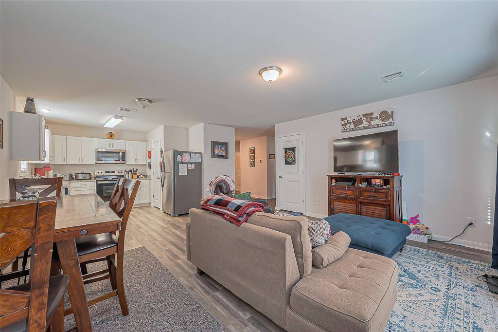This photo shows an open-concept living area with a modern kitchen and dining space. It features light wood flooring, a cozy beige sectional sofa, and a large TV. The kitchen has white cabinets, stainless steel appliances, and a dining table with wooden chairs, creating a welcoming and functional living space.