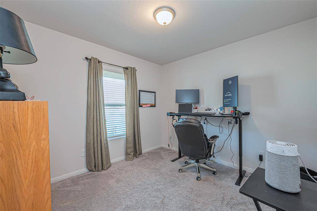 This room is set up as a home office with a desk, chair, and computer setup. It features a window with curtains, carpeted flooring, and provides a functional workspace with natural light.