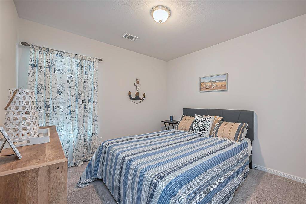 This cozy bedroom features a nautical theme with striped bedding and beach-inspired decor. It has a soft carpet, a small side table, and a dresser. A window with patterned curtains adds natural light to the space.