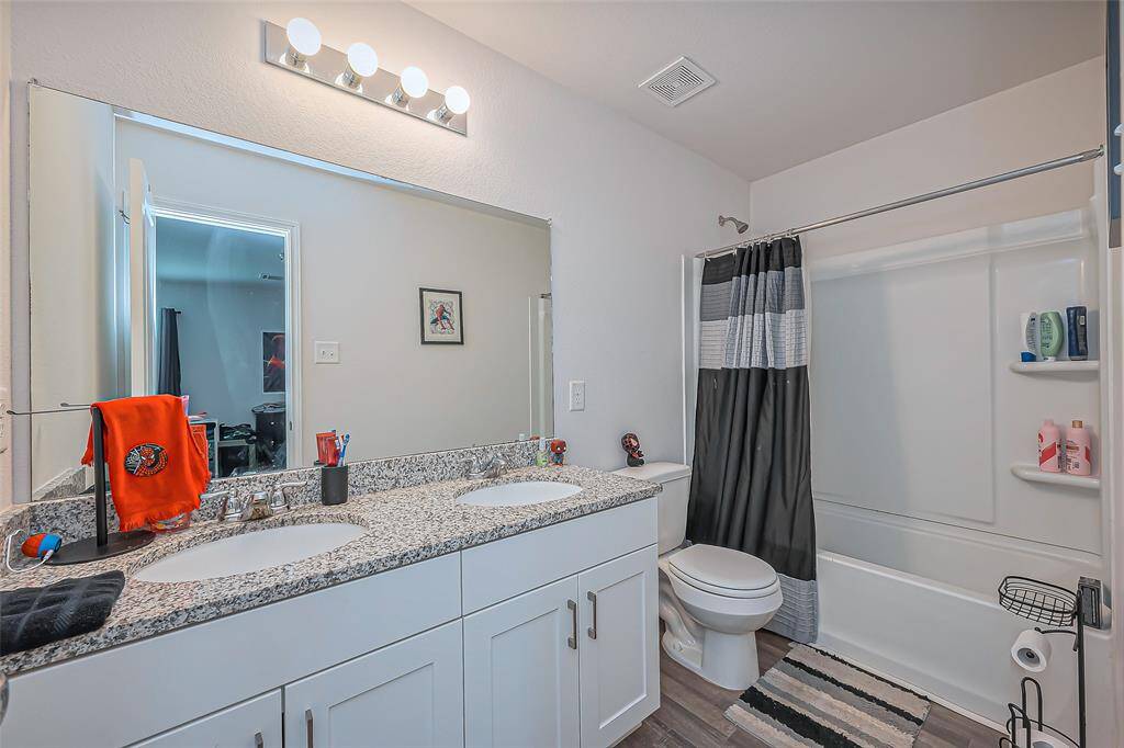 This bathroom features a modern design with a dual-sink granite countertop, large mirror, and a tub-shower combo with shelves. The space is well-lit and includes a sleek, contemporary vanity with ample storage.