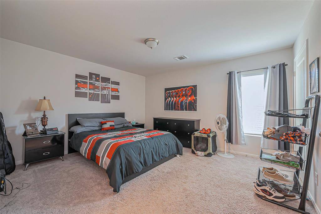 Spacious bedroom with neutral walls and carpet. Features a large bed, modern decor, and ample natural light from a window. Includes a dresser and shelving for storage, and a cozy atmosphere perfect for relaxation.