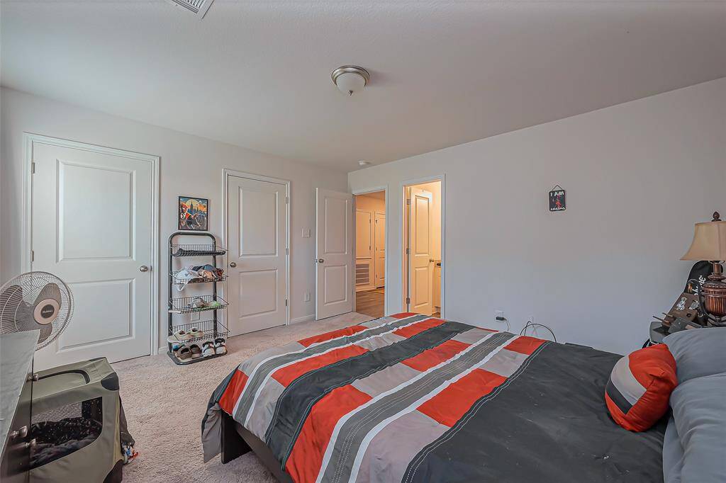 This is a cozy bedroom featuring neutral walls, carpeted flooring, and a simple ceiling light fixture. The room includes a bed with a colorful striped bedspread, a small shelf unit, and two doors leading to additional spaces. The overall atmosphere is inviting and functional.