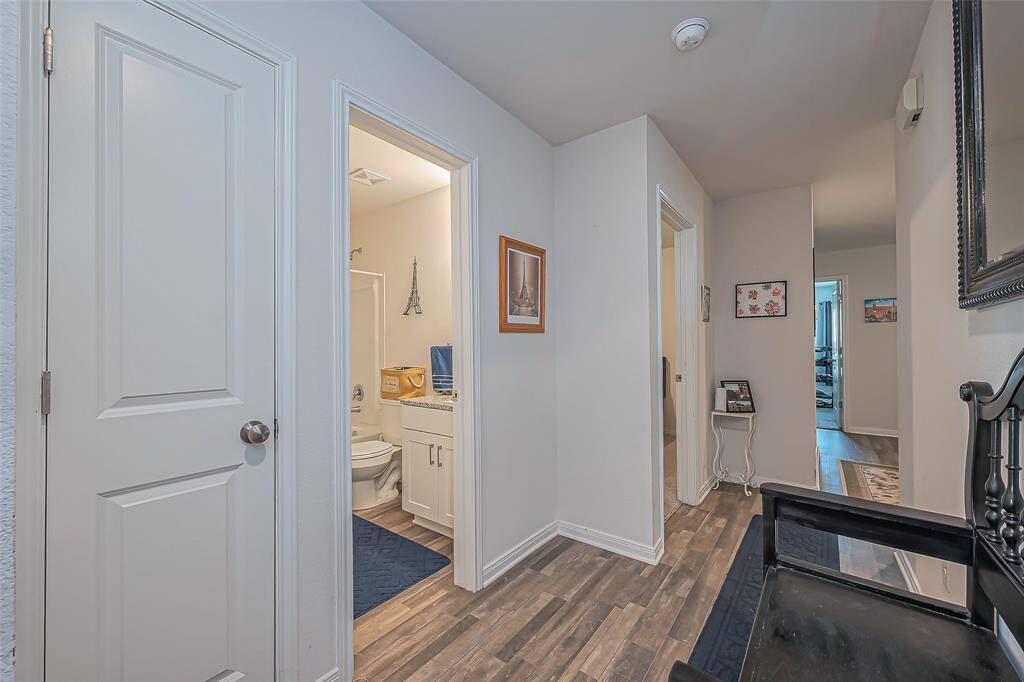 This photo showcases a hallway with wood-style flooring and white walls. It features a bathroom on the left with modern fixtures, a decorative mirror on the right, and leads to additional rooms, suggesting a functional layout with easy access to different areas of the home.