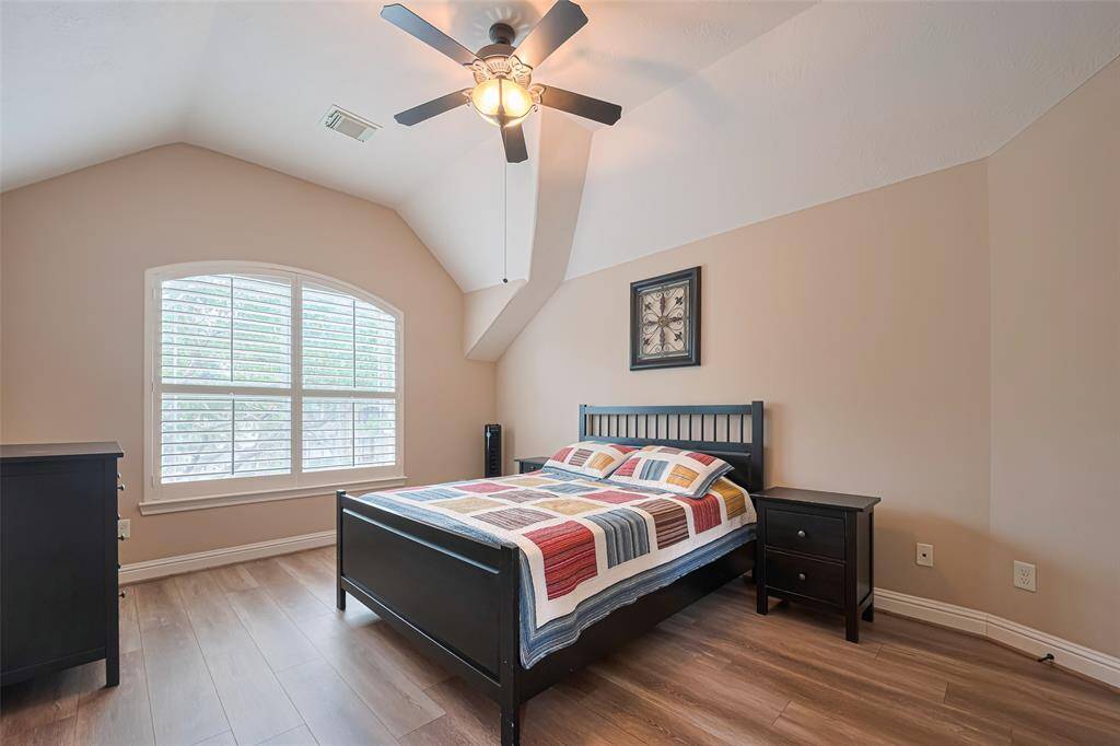 Another elegant and cozy bedroom showcases a vaulted ceiling, rich wood-style floors, and a large arched window fitted with plantation shutters for natural light