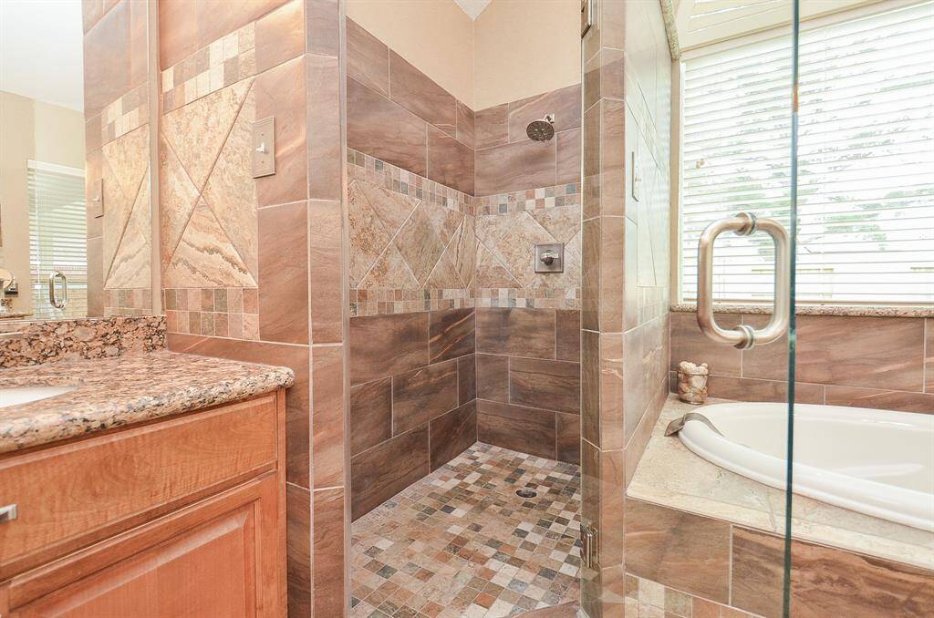 The premier bathroom features a spacious walk-in shower with intricate tile work, a granite countertop, and a large bathtub with a glass enclosure. Natural light flows through the window, creating a warm and inviting space.