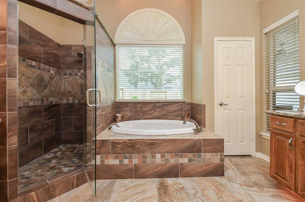 This elegant bathroom features a spacious glass-enclosed shower with rich brown tiles and mosaic accents. It includes a luxurious bathtub under a large window with a half-circle design, allowing ample natural light.