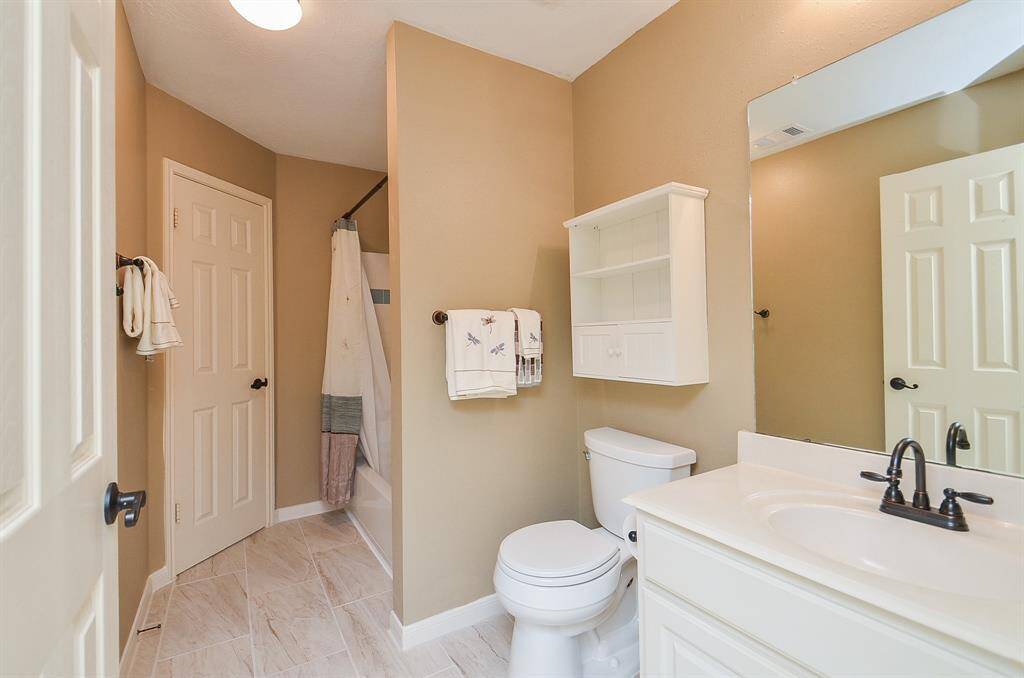 This bathroom features a clean, modern design with neutral tones. It includes a bathtub/shower combo, a white vanity with a large mirror, and a toilet. The floor is tiled, and there is a small wall cabinet for storage.