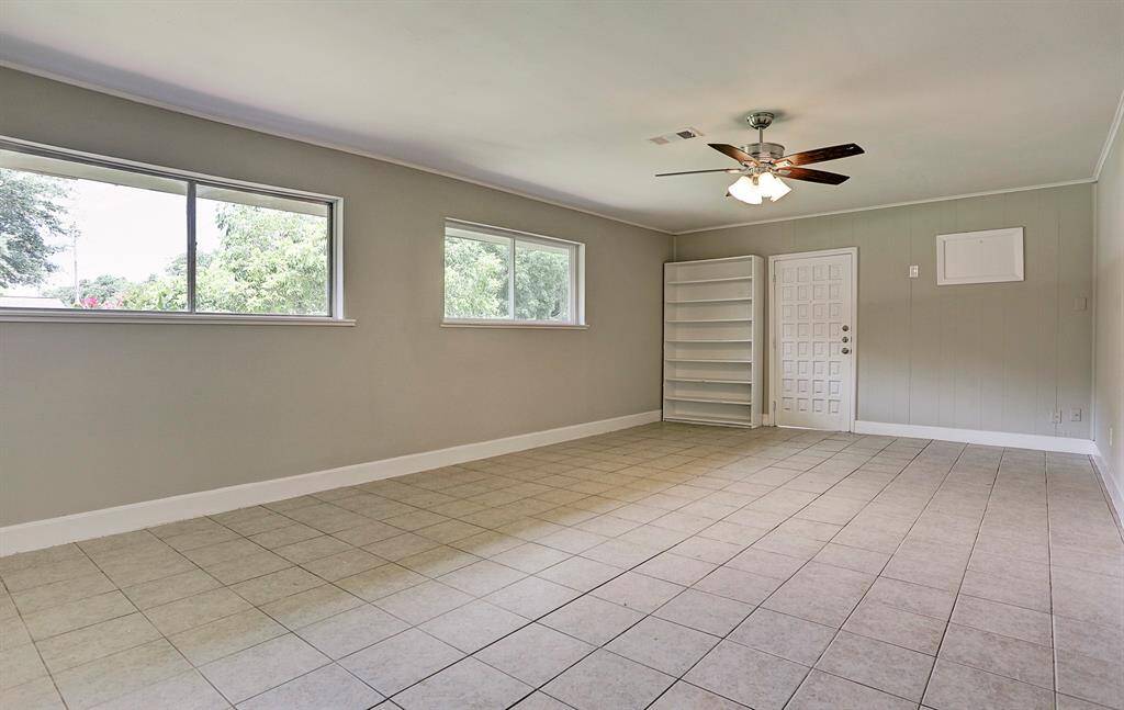 This spacious room features large windows providing ample natural light, neutral tile flooring, light gray walls, a ceiling fan, and built-in shelving, offering versatile use for living or office space.