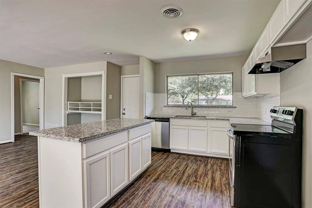 This kitchen features a modern design with granite countertops, white cabinetry, stainless steel appliances, and wood-style flooring. A large window provides ample natural light, and the open layout flows into an adjacent room. Perfect for cooking and entertaining.