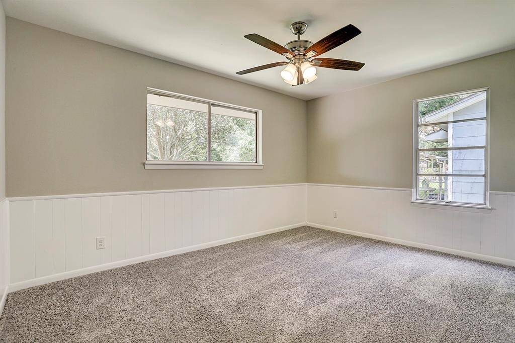 This room features neutral walls with white wainscoting, plush carpet, and a ceiling fan with lights. It has two windows providing natural light, creating a bright and airy atmosphere.
