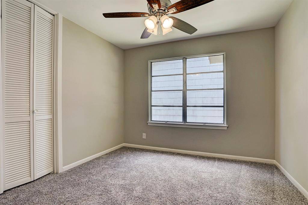 This cozy bedroom features soft carpeting, neutral walls, a ceiling fan with lights, and a large window for natural light. A louvered closet door provides storage space.