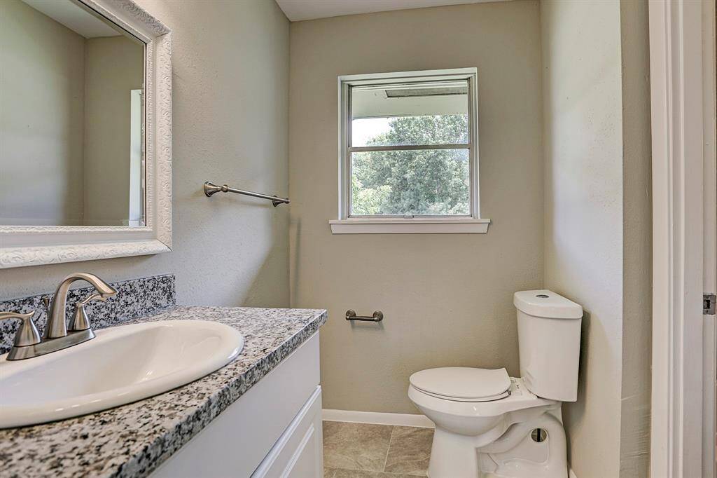 This is a compact bathroom featuring a granite countertop with an undermount sink and modern faucet. It has a small window for natural light, a mounted mirror, and a toilet, all set against neutral-colored walls.
