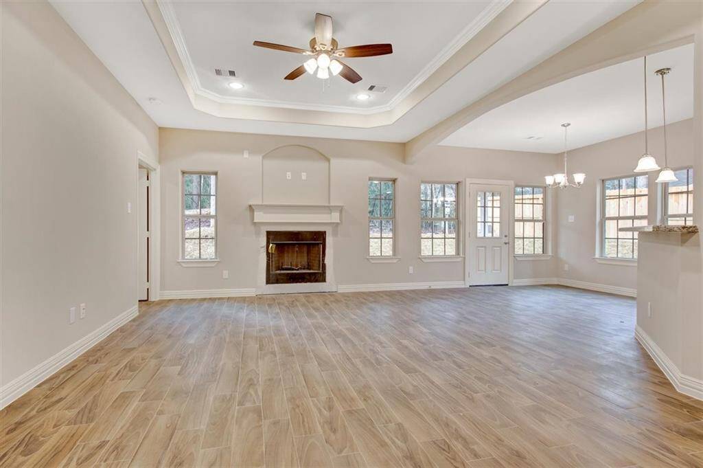 This spacious living area features a cozy fireplace, tray ceiling with fan, and abundant natural light from multiple windows. The open layout flows into a dining space, highlighted by elegant pendant lighting and wood-look flooring.