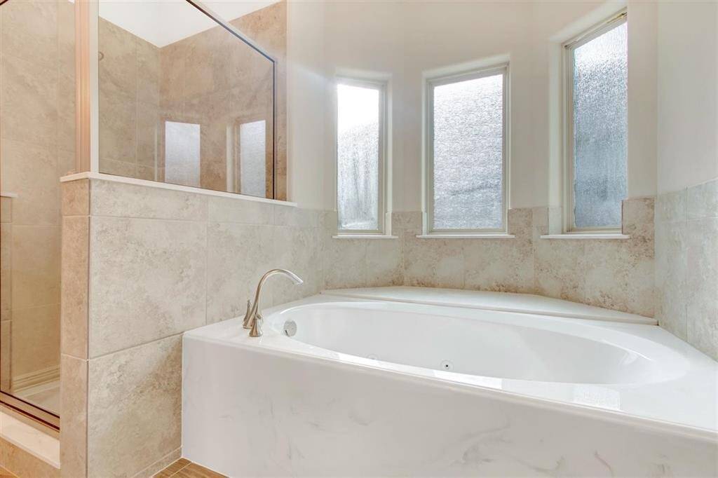 Spacious bathroom featuring a large soaking tub with elegant tiling, adjacent to a glass-enclosed shower. Natural light streams in through three frosted windows, creating a bright and inviting atmosphere.