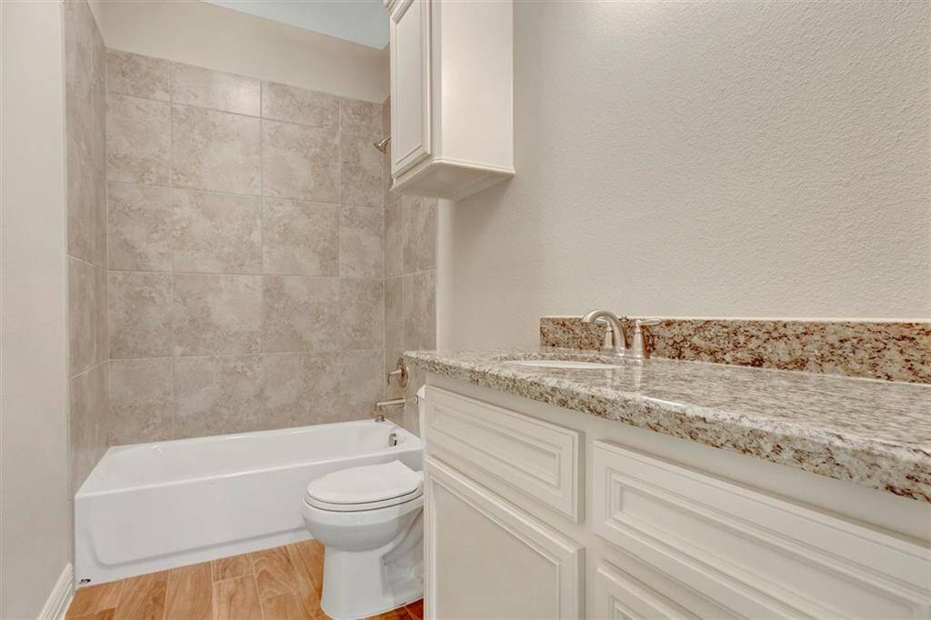 This bathroom features a modern design with a granite countertop, white cabinetry, and a bathtub with tiled surround. The floor is finished with wood-like tiles, offering a clean and elegant look.
