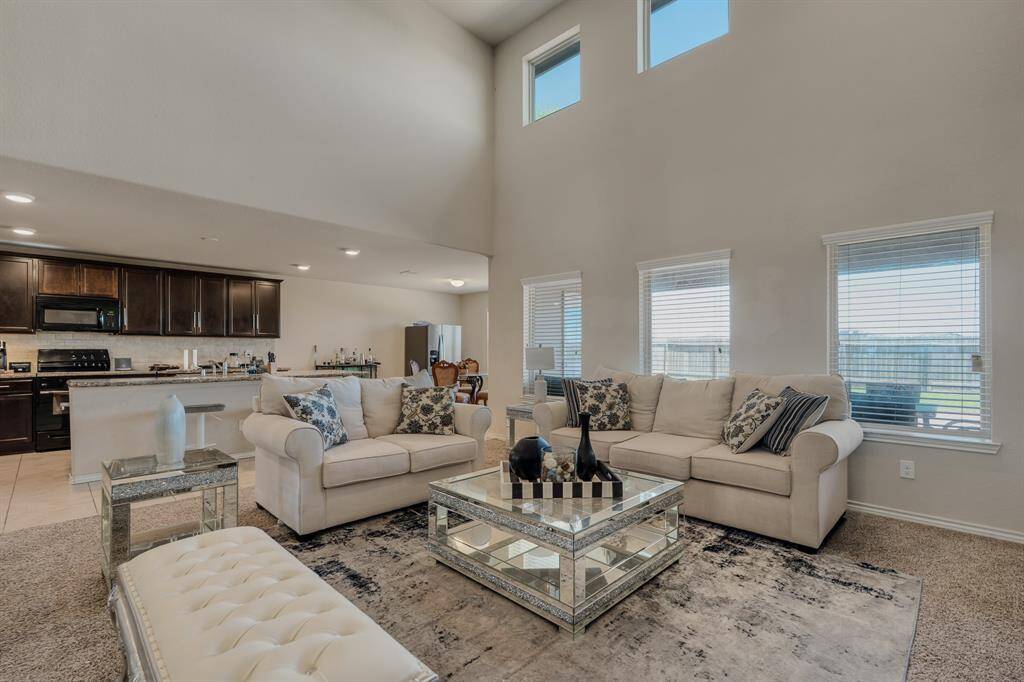 Open-concept living area features high ceilings and abundant natural light. It includes a modern kitchen with dark cabinetry, a cozy seating area with plush sofas, and stylish decor, perfect for comfortable living and entertaining.