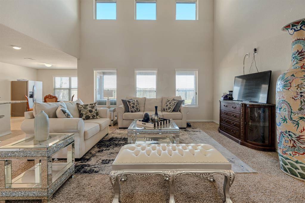 Living room with high ceilings and large windows, offering plenty of natural light. Features elegant furnishings, including plush sofas and a decorative vase, along with a mounted TV. Open layout connects to the kitchen and dining area.