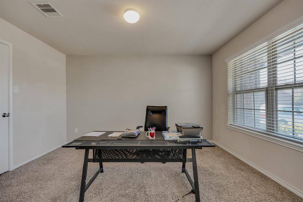 This room offers a bright, inviting space perfect for a home office, featuring a large window for natural light, neutral walls, and carpeted flooring.