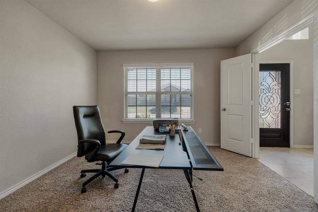 Home office with a large window, neutral walls, and carpeted flooring. A sleek desk and chair are positioned near the window, and a distinctive front door with decorative glass is visible.