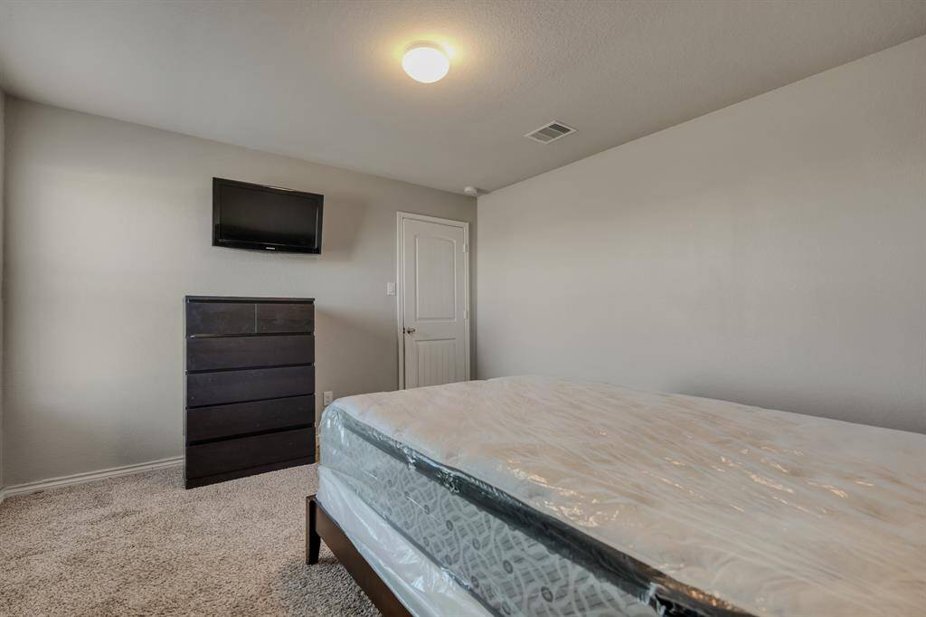 This is a simple bedroom featuring neutral walls, carpeted flooring, a ceiling light, a mounted TV, and a dark dresser. The room also includes a bed with a new mattress, still in protective plastic.