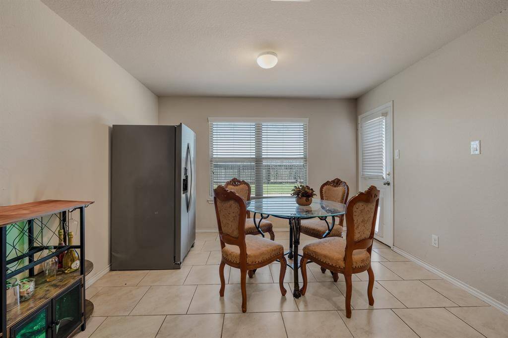 This cozy breakfast area features a round glass table with four elegant chairs, a large window for natural light, and a stainless steel refrigerator. The space is tiled and has a neutral color palette, offering a bright and inviting atmosphere.