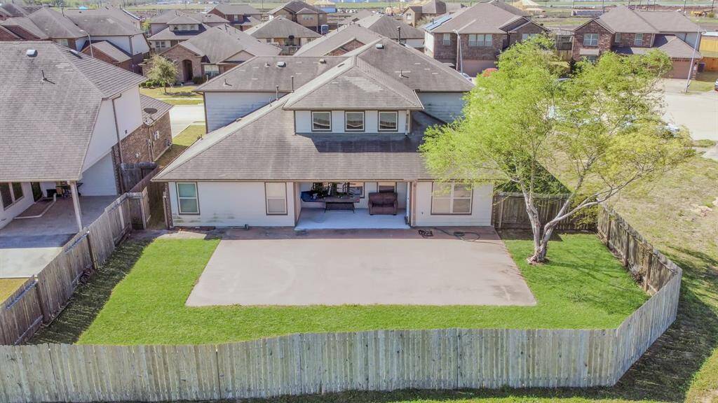 A two-story home with a fenced backyard featuring a large patio area and a well-maintained lawn. A mature tree provides shade on one side, and neighboring houses are visible nearby.
