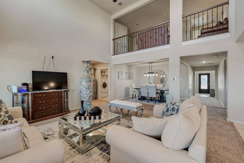 Spacious living area with high ceilings and an open layout. Features a cozy seating area, elegant dining space, and a visible upper loft. Neutral tones and ample natural light create a welcoming atmosphere.