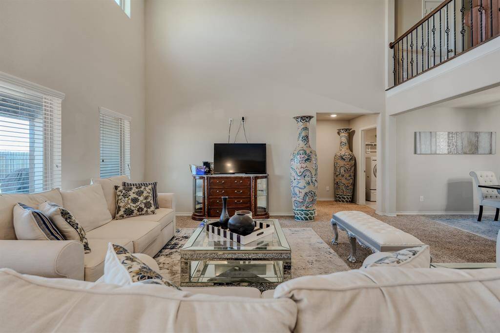 High ceilings, ample natural light from large windows, and elegant décor. It includes cozy seating, a glass coffee table, and a TV setup. Decorative vases add a touch of sophistication, and an open layout connects to an adjacent area, enhancing the airy feel.