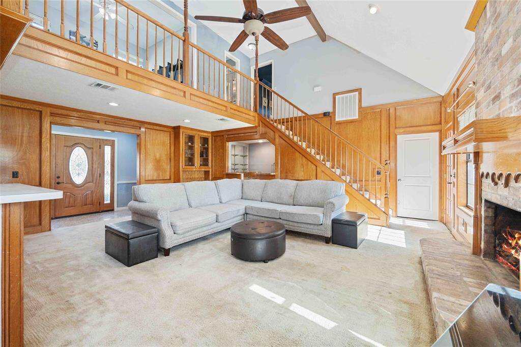 This inviting living room features high ceilings with wooden beams, a cozy fireplace, and an open staircase leading to a loft area. The space is bright and airy, with a large sectional sofa and a ceiling fan for comfort. Wood paneling adds warmth, while the front door with glass accents enhances natural light.